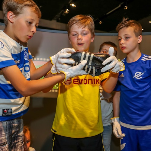 三個穿著足球球衣和白色手套的男孩在一個像博物館的環境中檢查一隻鞋子。