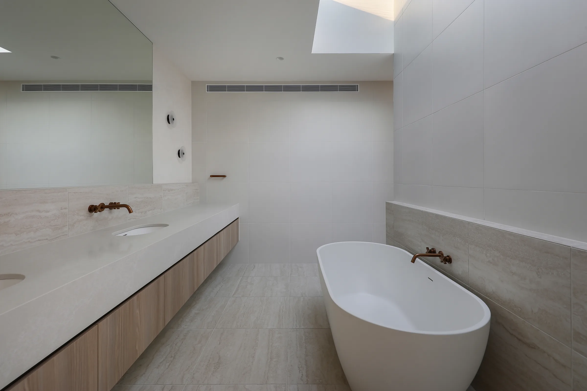 Main bathroom with travertine-look tiles, twin undermount basins on a stone vanity, a freestanding white bath, copper tapware and a skylight