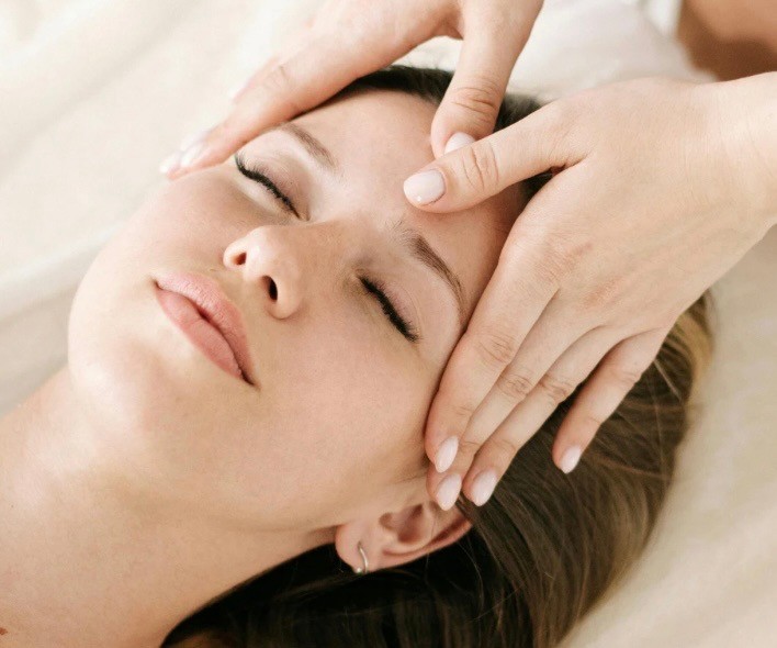A woman lying down in a spa setting, relaxing with her eyes closed. Sunlight streams through blinds, casting striped shadows on her face. The scene conveys a sense of serenity and well-being.