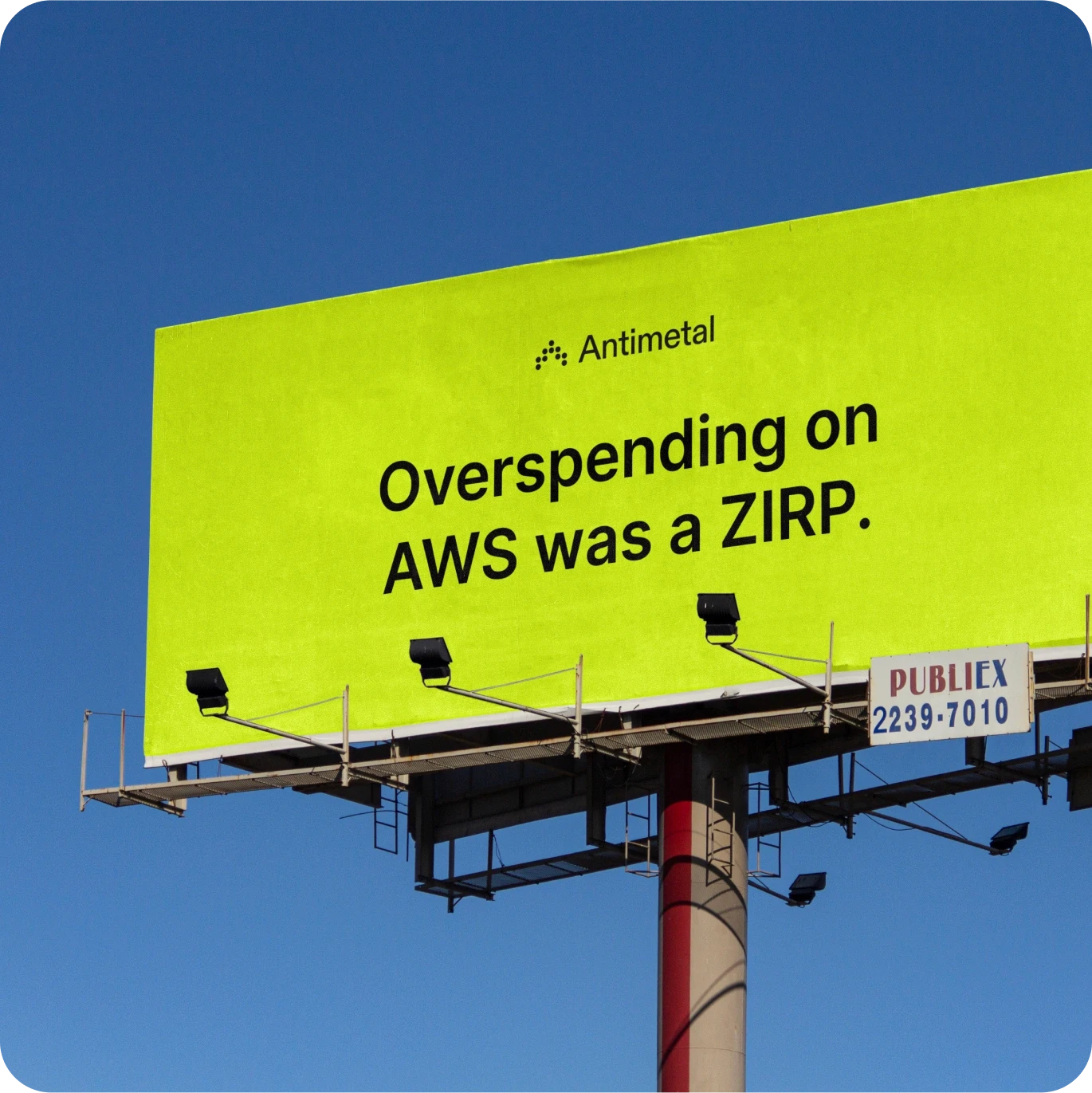 A bright neon yellow billboard against a clear blue sky features the Antimetal logo and the text "Overspending on AWS was a ZIRP." The simple design with bold black letters aims to draw attention to AWS cost management. The billboard is mounted on a metal structure with floodlights and a small sign from the advertising company PUBLIEX.