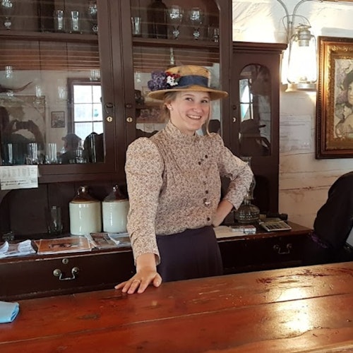 Uma mulher em trajes históricos está atrás de um balcão de madeira, sorrindo. Prateleiras com potes e decoração vintage estão visíveis ao fundo.