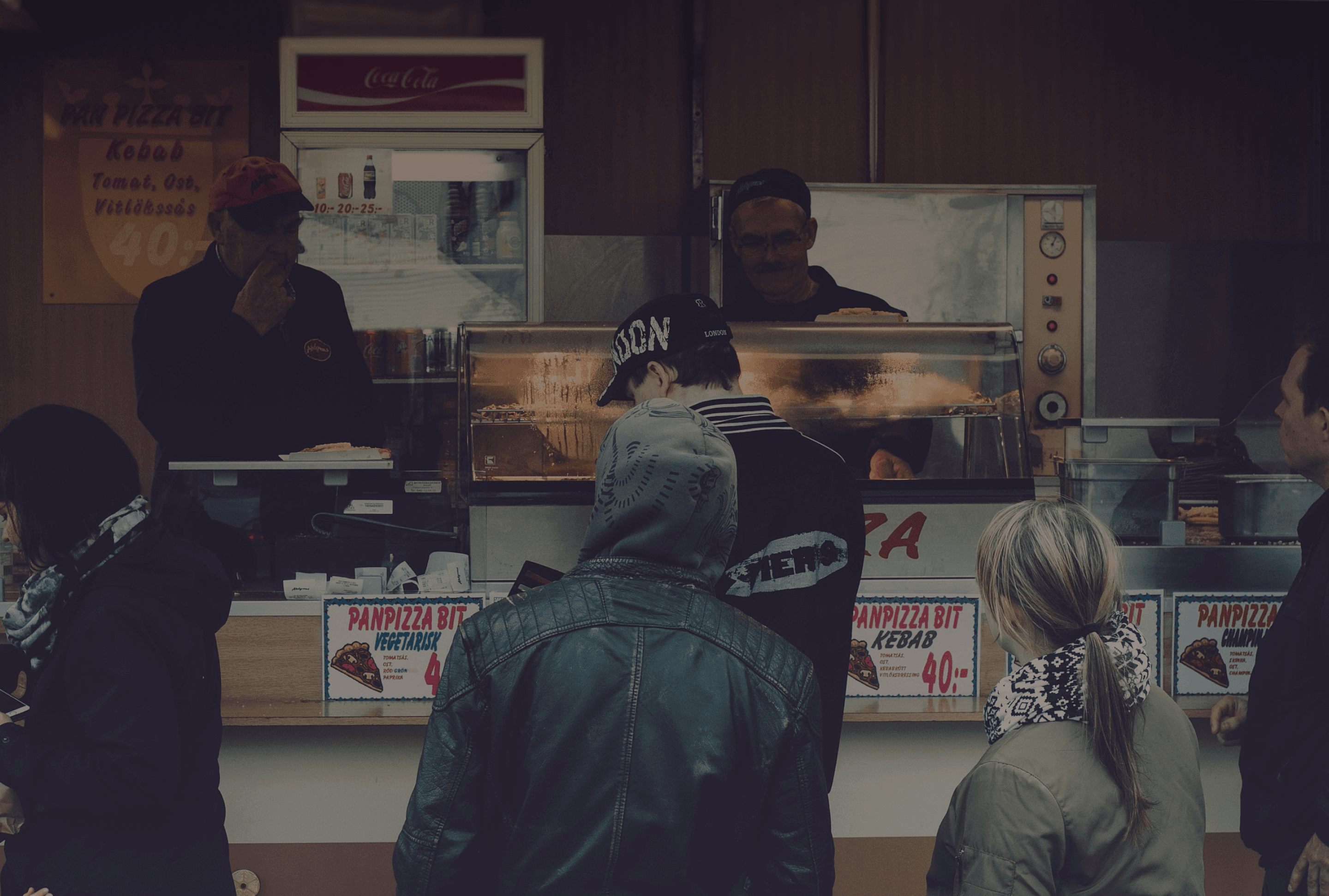 Un grupo de personas haciendo fila en un puesto de comida o pequeño restaurante. Los clientes parecen estar esperando para hacer o recibir sus pedidos, mientras que los trabajadores son visibles detrás del mostrador, preparando la comida.