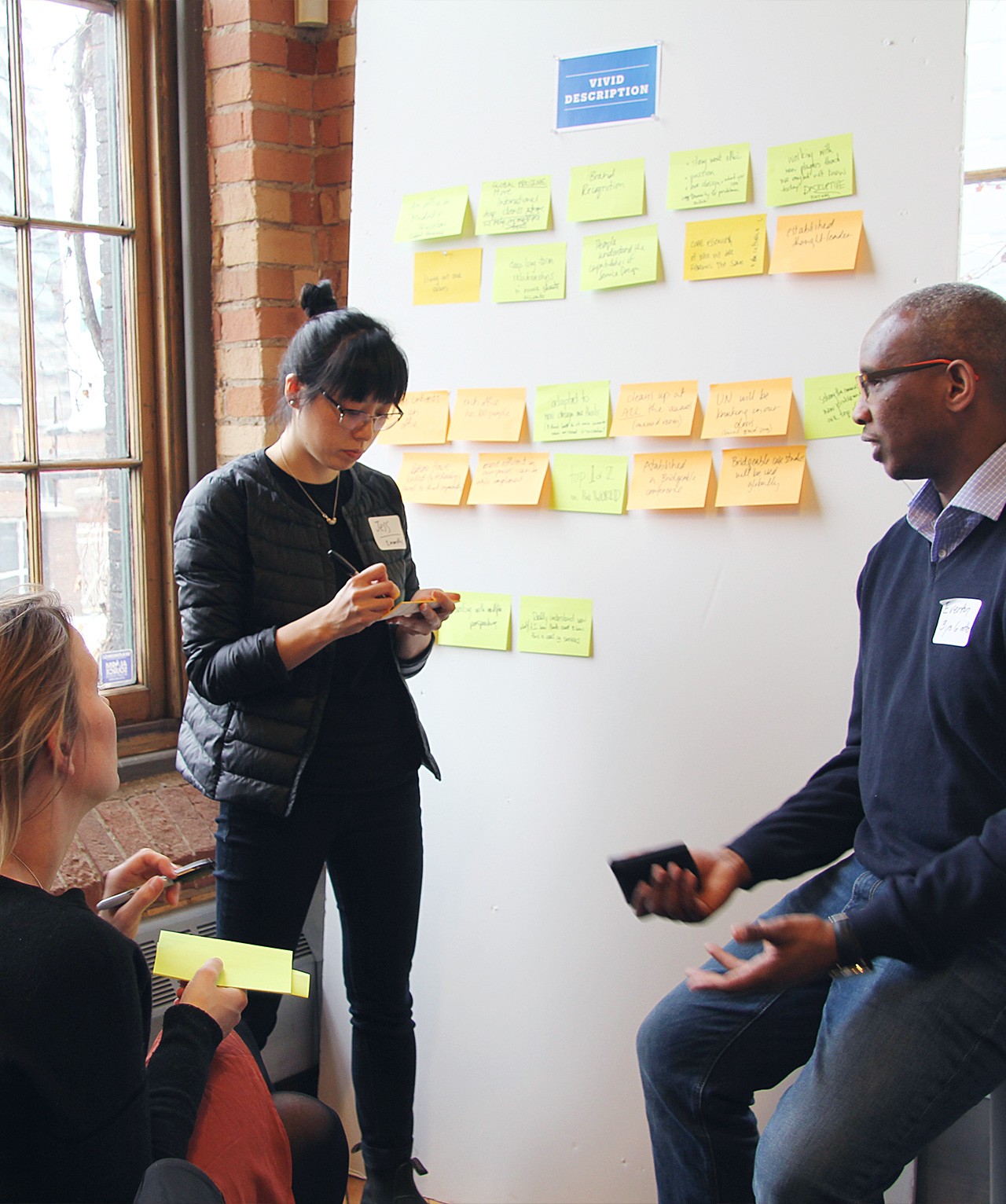 Three people standing around a wall with post-it notes. One person is taking notes.