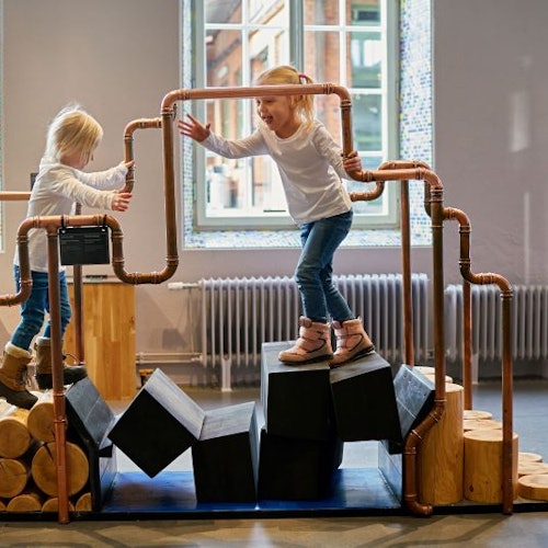 Two children play on a climbing structure made of copper pipes with wooden logs and black blocks, indoors near a window.
