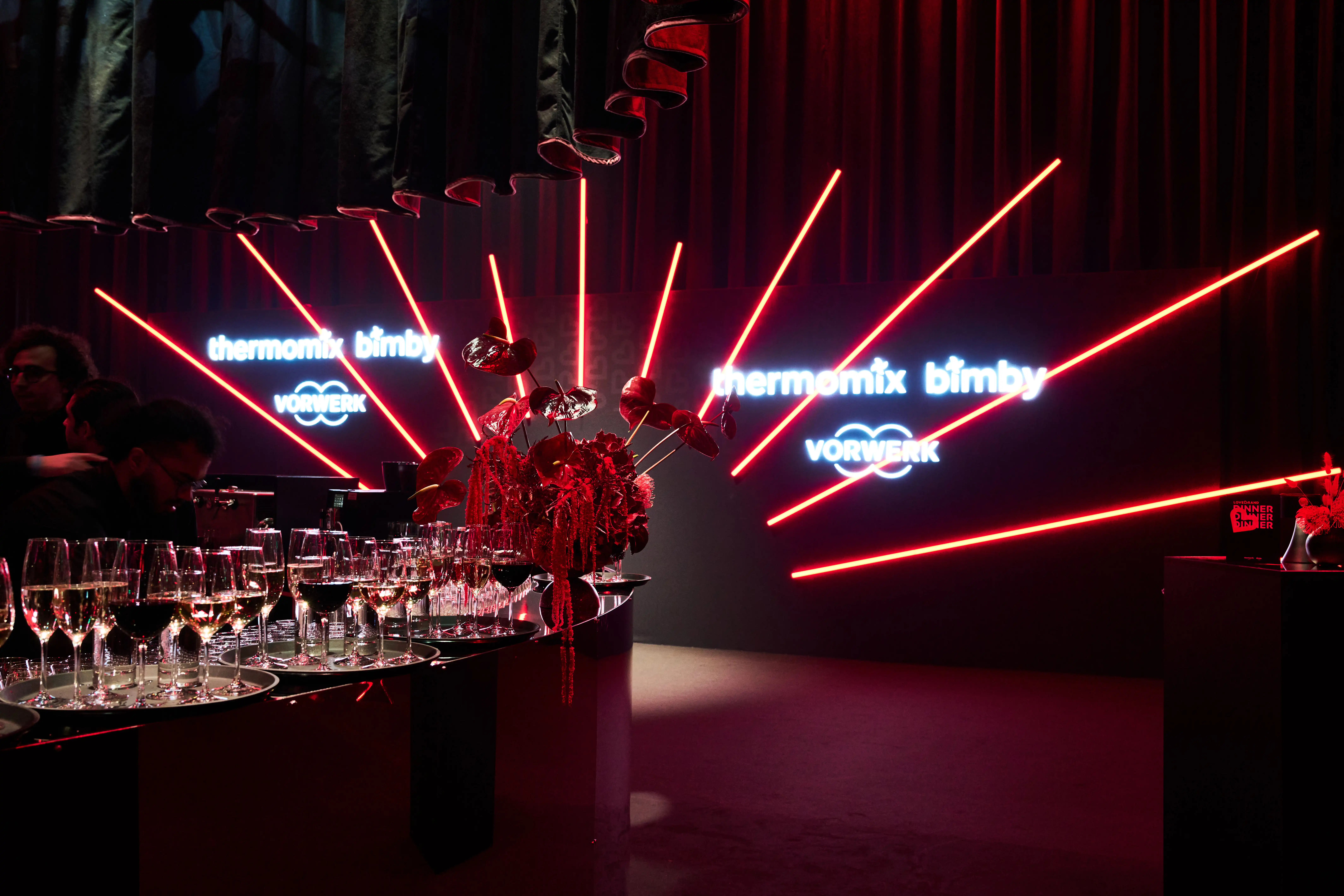 Bar with rows of wine glasses in front of neon Thermomix Bimby signs and red light rays.