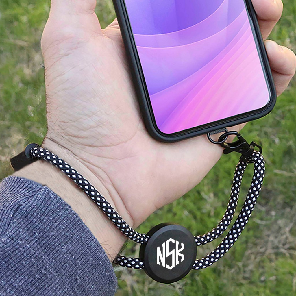 A customizable phone holder wristlet being worn on someones wrist connected to a phone. All you can see is a persons hand hovering over grass holding the phone.