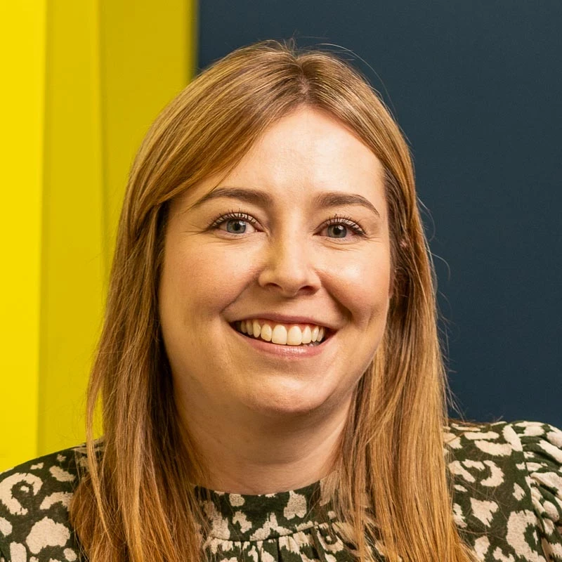 Person with straight, light brown hair wearing a patterned top, standing indoors against a background with yellow and dark blue panels.