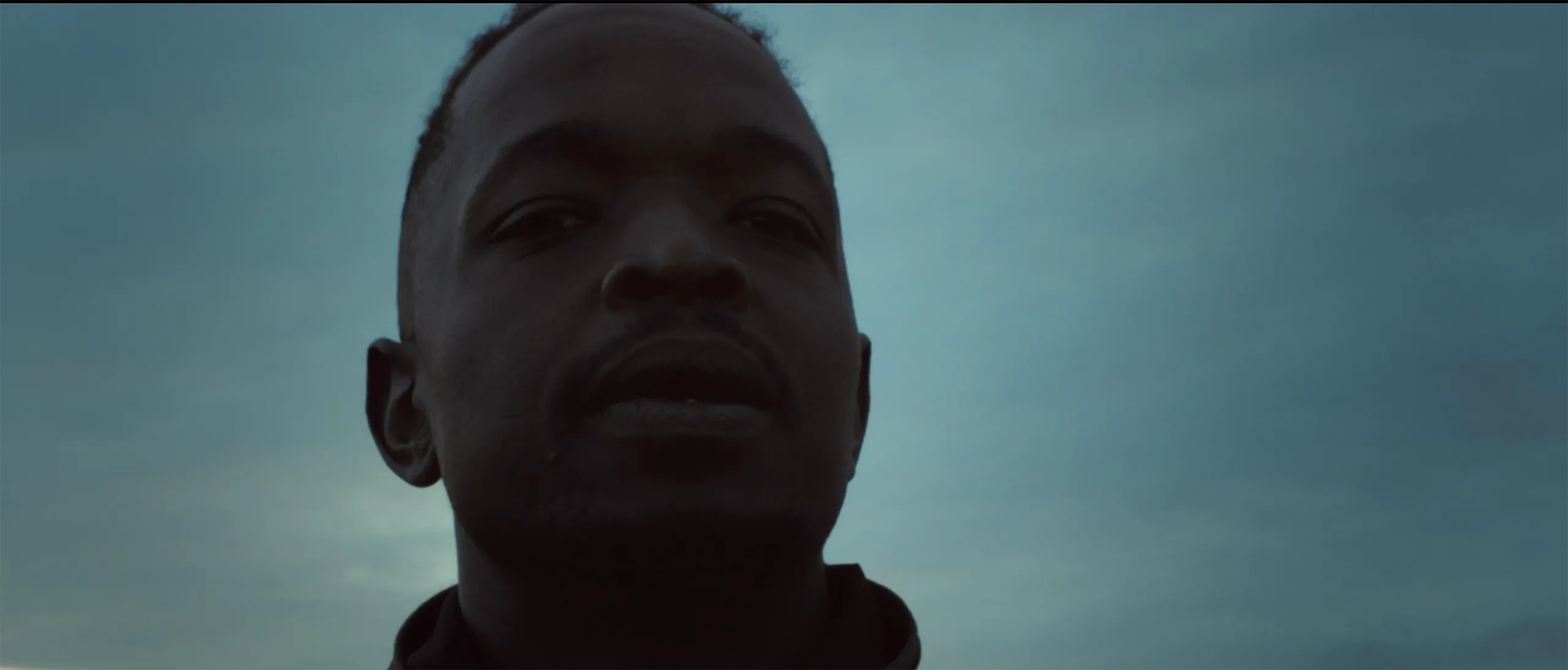 A low angle cinematic portrait of a man looking upward against a moody desaturated overcast sky.