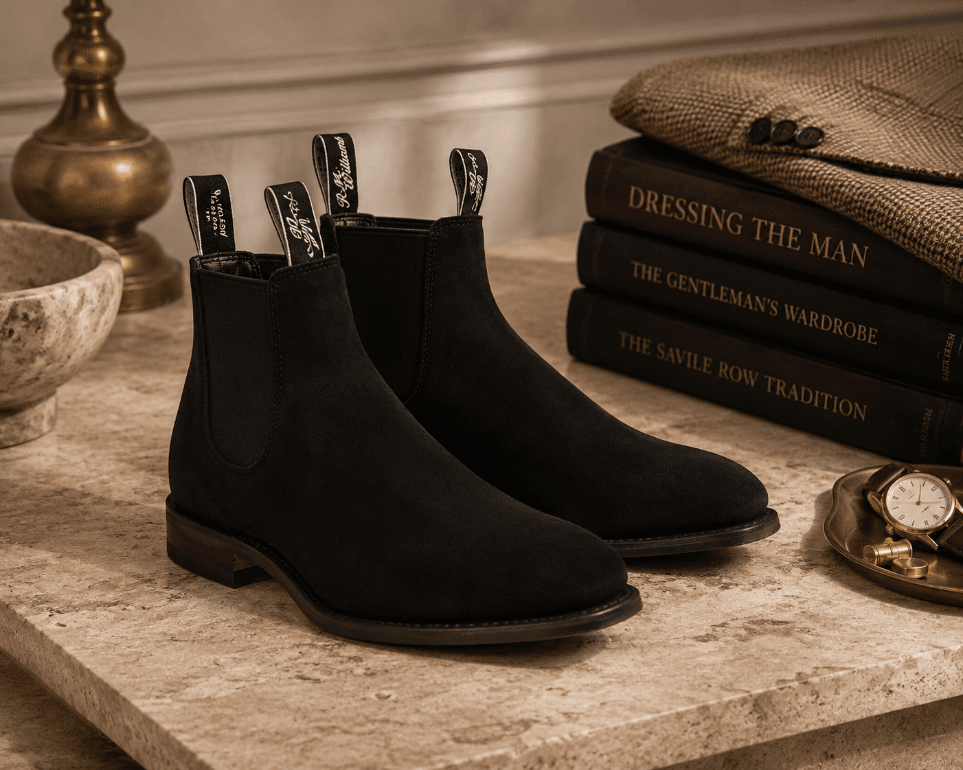 A pair of black suede RM Williams Chelsea boots featuring signature branded pull tabs sitting on a travertine stone ledge. The refined menswear setup includes stacked hardcover books titled "Dressing the Man", "The Gentleman's Wardrobe", and "The Savile Row Tradition", topped with a patterned tweed jacket, alongside a brass lamp, a stone bowl, and a valet dish with a gold watch under warm lighting.