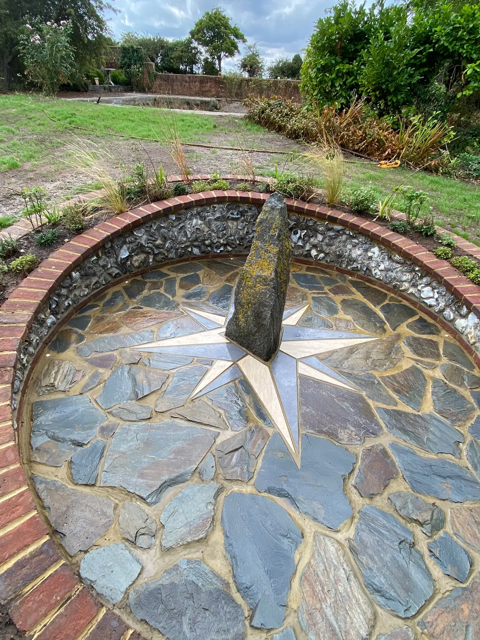A circular stone fountain with a central pillar, surrounded by greenery and blue skies above.