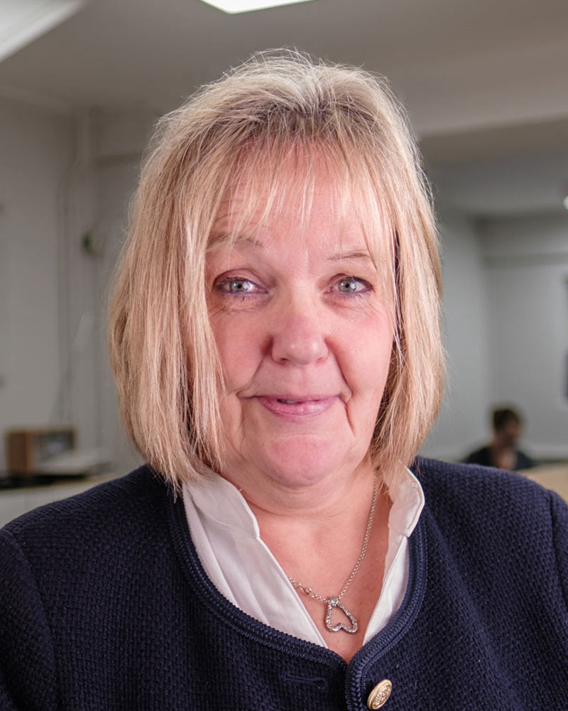 A portrait of Tina, a receptionist at Cricklade Dental Practice, smiling and wearing a white blouse under a dark blue textured jacket with gold buttons. She is wearing a silver heart-shaped necklace and is standing in the practice reception area with the practice logo visible on a glass partition in the background.