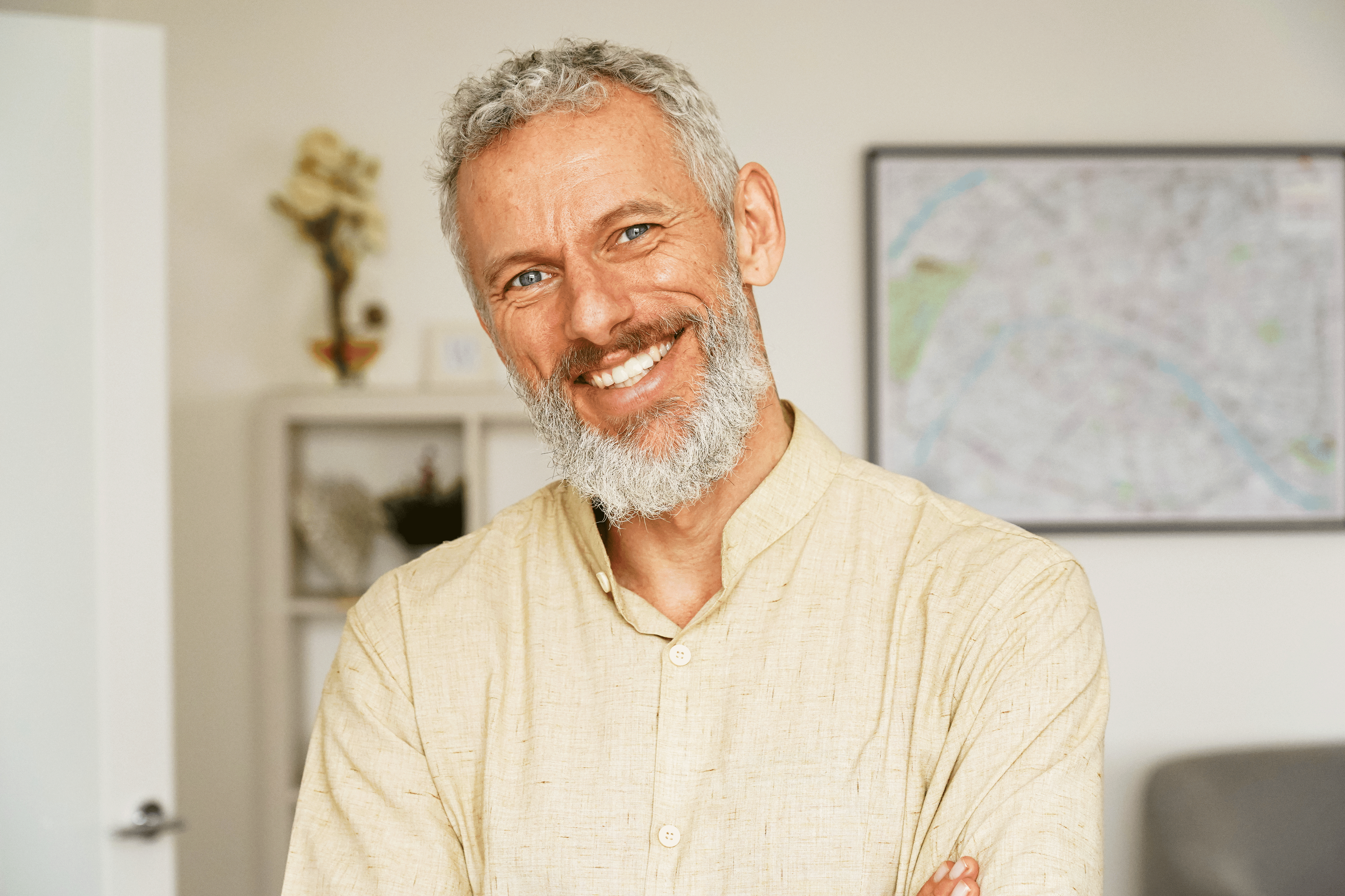 A smiling man with short, curly grey hair and a full grey beard looks directly at the camera with his arms crossed. He is wearing a light-colored, button-down shirt and is standing in a brightly lit room with a framed map visible in the background.