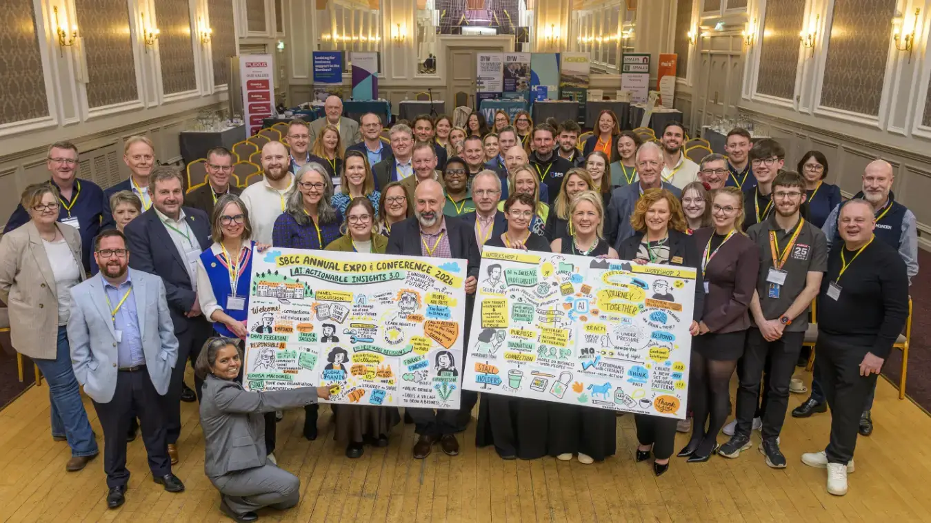 A large group of people at a conference pose with illustrated boards featuring diagrams and text. The room is ornate, with chandeliers and banners. Smiling faces create an energetic, collaborative atmosphere.