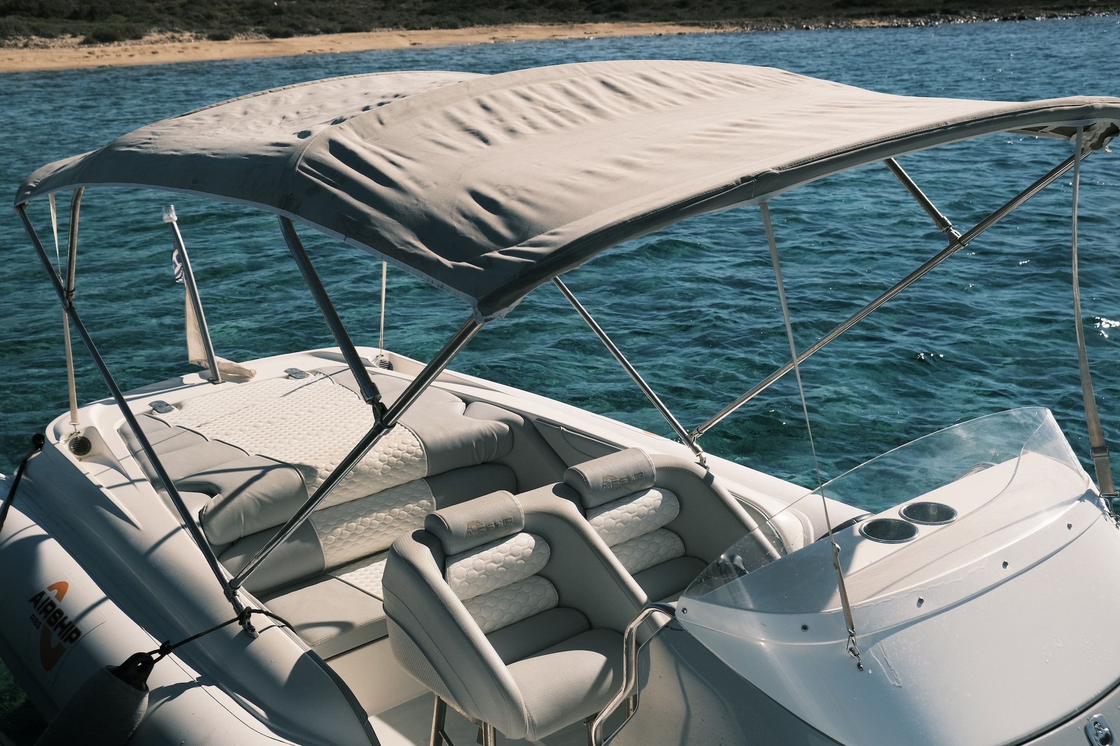 White luxury yacht bow with cushioned seating area anchored in turquoise waters near a sandy beach with green hills in the Cyclades.