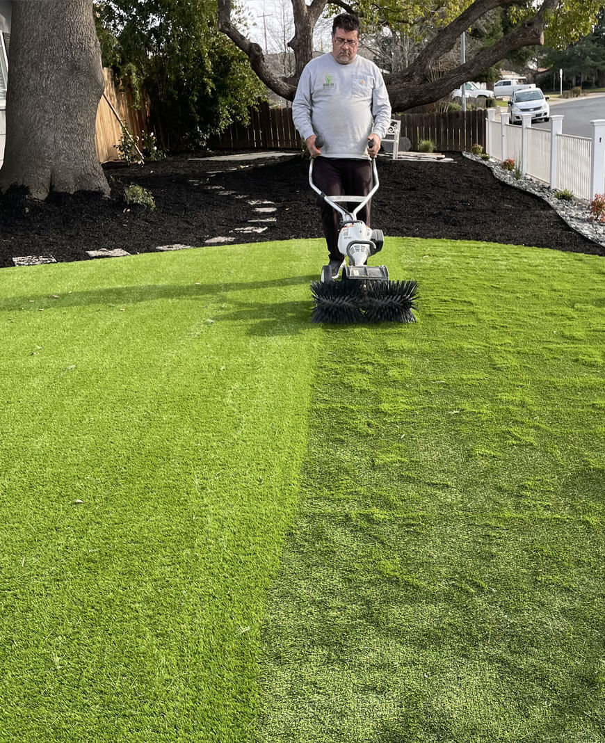 On a beautiful sunny day, an AGL Grass North installer pushes a power broom across a fresh residential installation of AGL Grass artificial turf.