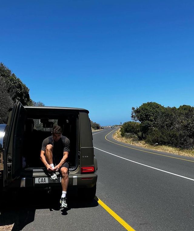 Focused young man in a mint green jacket staring ahead, with a fencing sword vertically aligned with his face, rooftops of a European city in the background.
