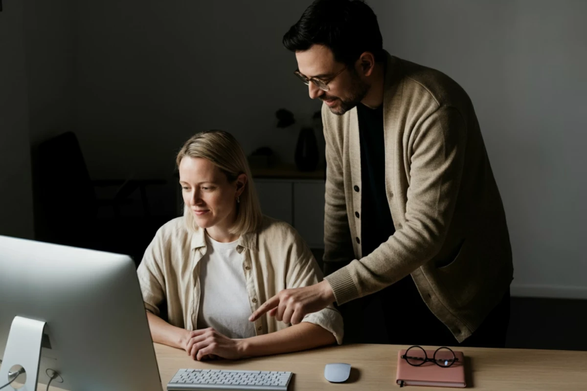 A man in a sweater leans over a woman at her desk, both engaged in a discussion, with a computer in view.