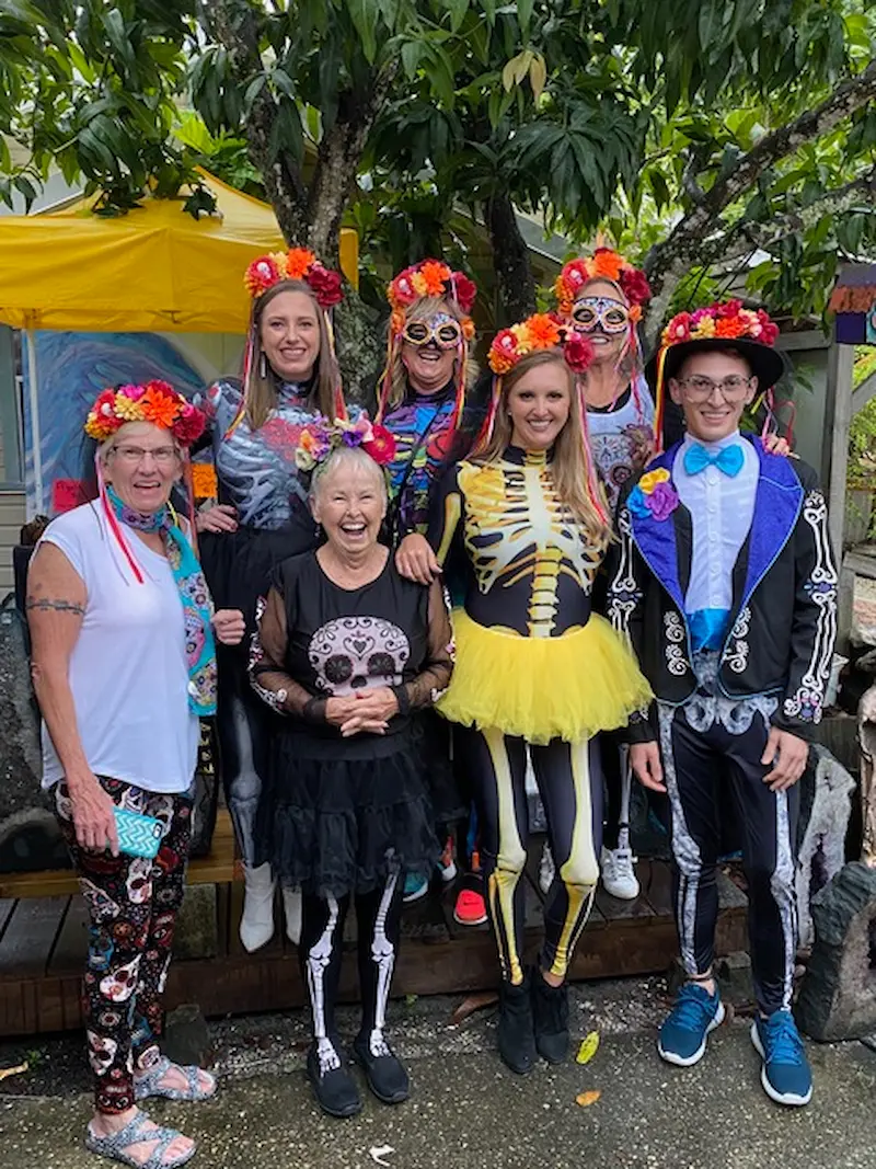 A group of people dressed in vibrant, intricately detailed Dia de los Muertos costumes and floral headpieces gather cheerfully in the Village of the Arts, Bradenton, FL, surrounded by lush greenery and a bright yellow tent in the background.