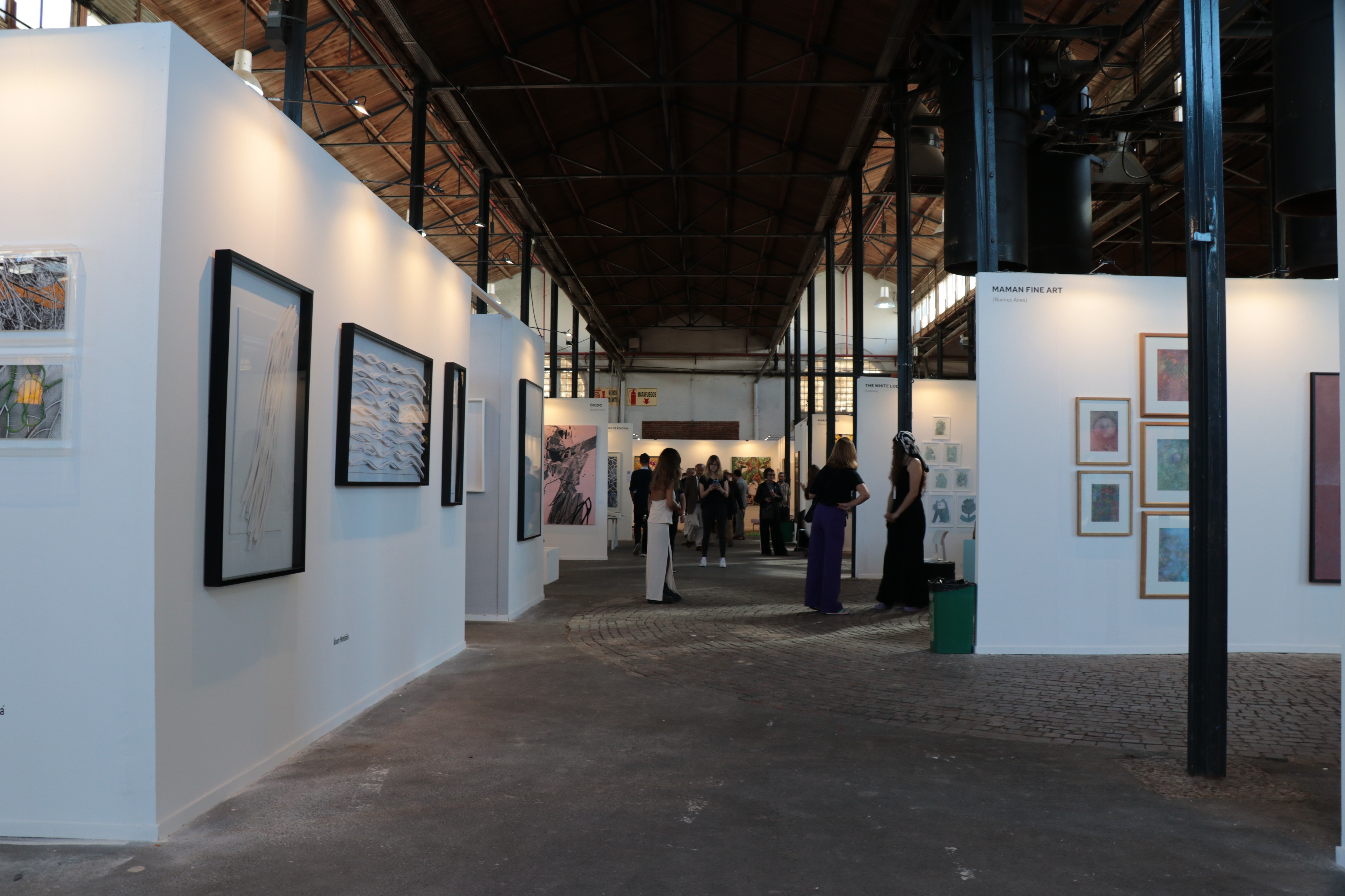 Esta foto captura el ambiente general del espacio de la feria MAPA durante el evento. El lugar, caracterizado por su arquitectura industrial, está lleno de asistentes que se mezclan y ven las obras de arte. La configuración incluye varias obras de arte exhibidas a lo largo de las paredes blancas de la galería, iluminadas por la iluminación del lugar, contribuyendo a un ambiente vibrante y atractivo.