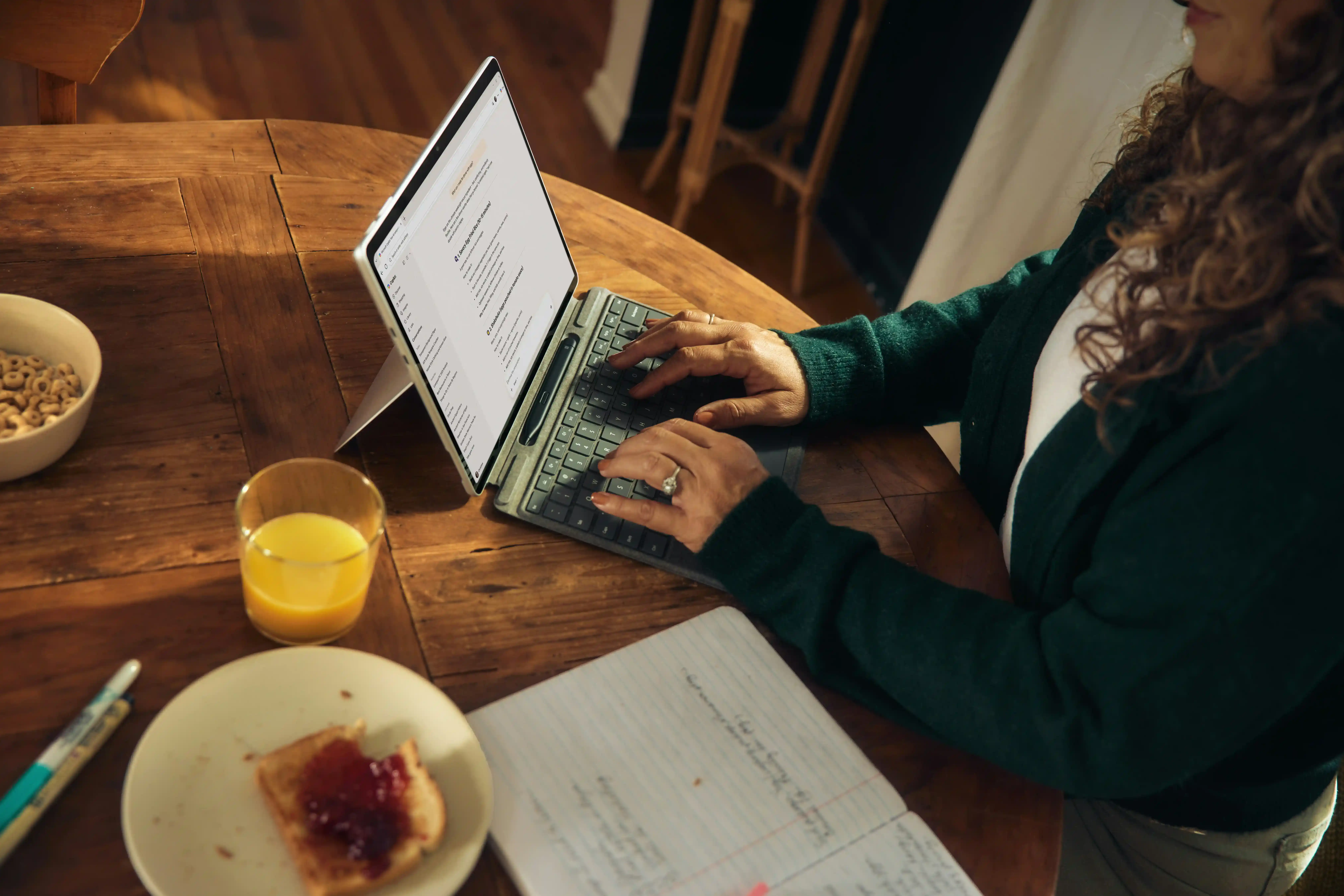 Person working on a laptop at a wooden table with documents, a glass of juice, and a plate of toast