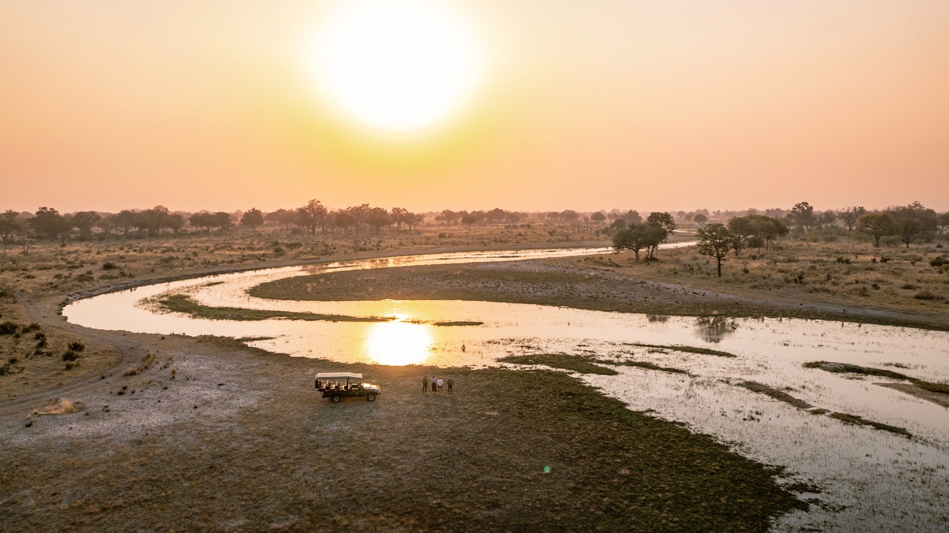 Kujwana Delta Ride, Okavango Delta, Botswana. Drönarvy över fyra personer på game drive blickar ut över solnedgången, med en safarijeep bredvid sig.