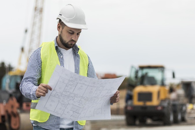 A construction worker in a hard hat and safety vest reviewing blueprints at a building site with machinery.
