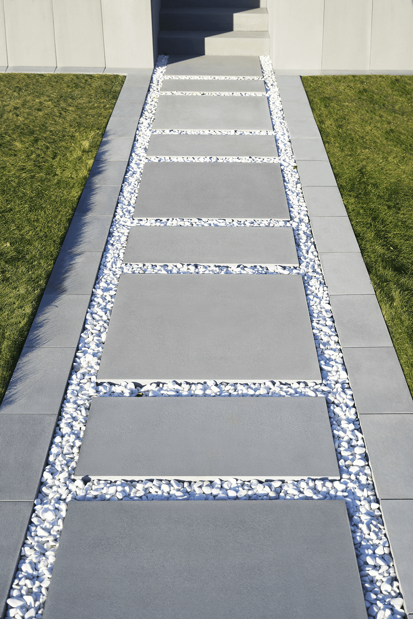 Moderní dům s velkými okny, kamennou cestičkou a upraveným trávníkem, obklopený stromy pod jasnou modrou oblohou.