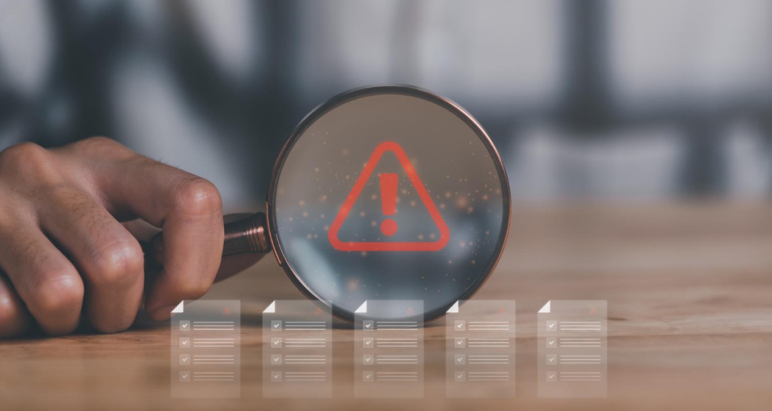 A close-up of a person holding a magnifying glass over a red warning icon with an exclamation mark, symbolizing risk or alert detection. Blurred digital checklists appear below the magnifying glass, representing monitoring or compliance review processes.