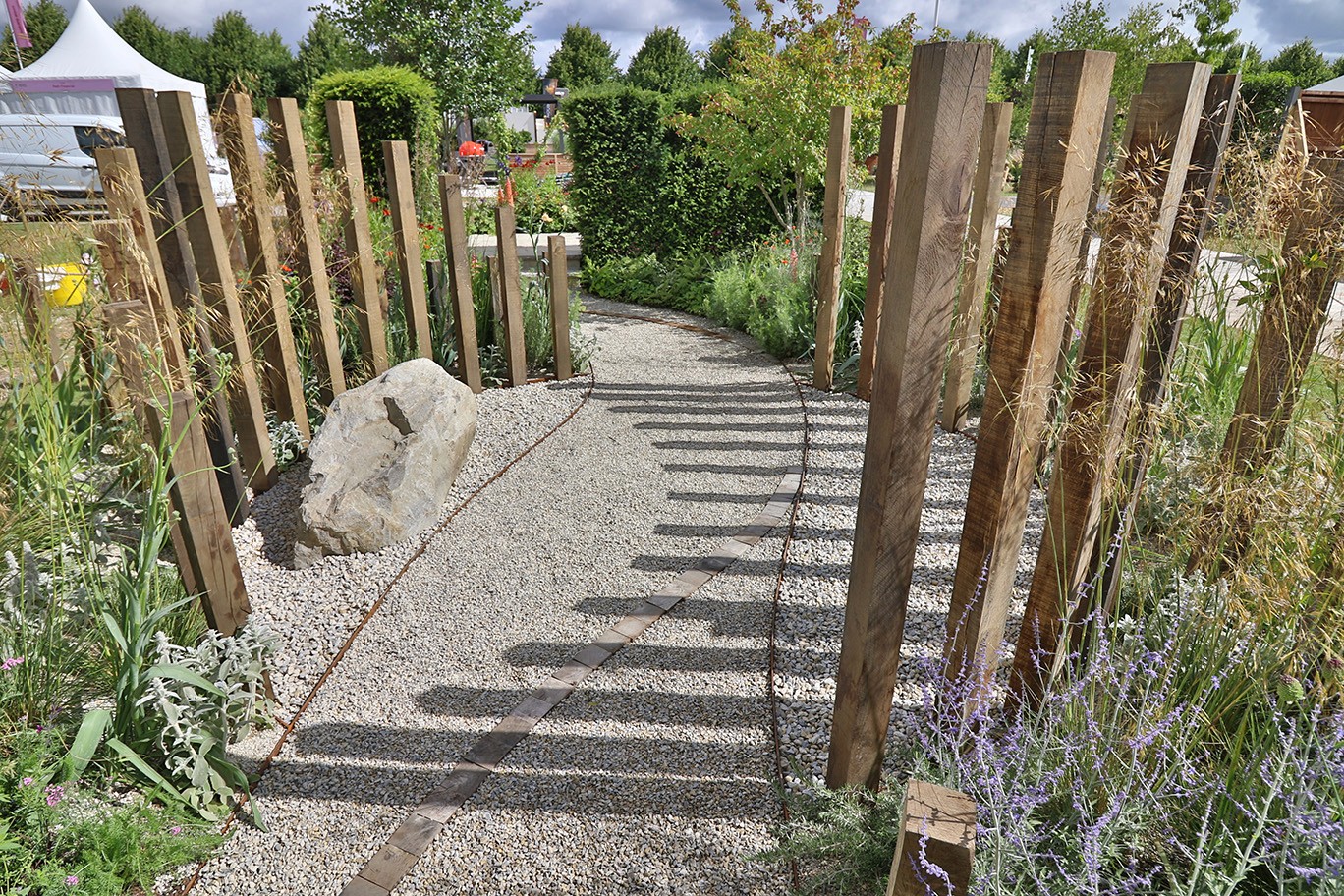 A wooden pathway leads through lush greenery, framed by vertical wooden posts, under a clear blue sky.