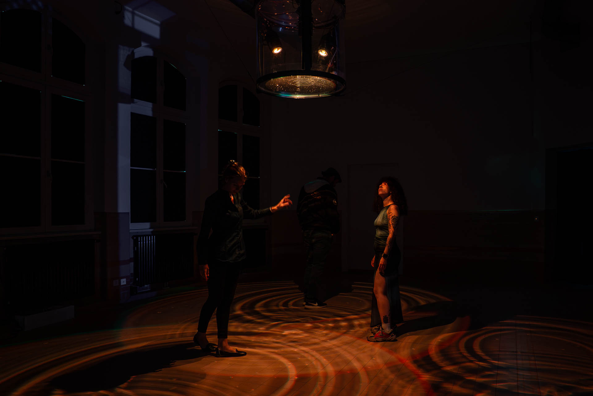 Three visitors in a dark room under a hanging glass cylinder that casts rippling amber light on the floor