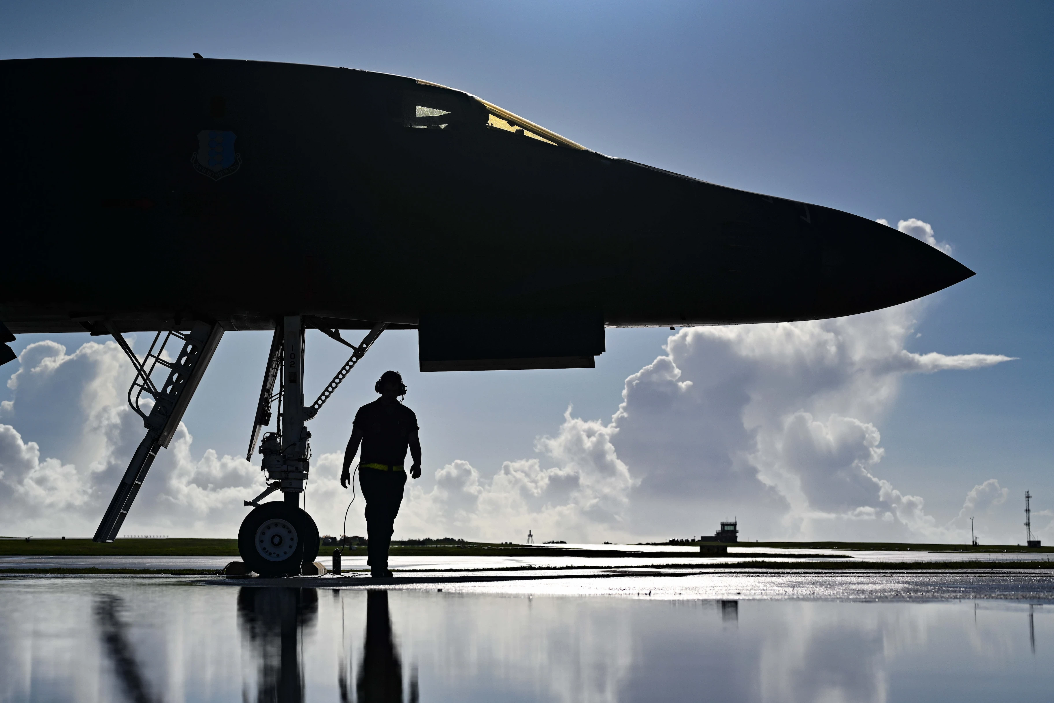 Silhouette of a ground crew member walking near the front landing gear of a military aircraft on a reflective wet runway under a cloudy sky.