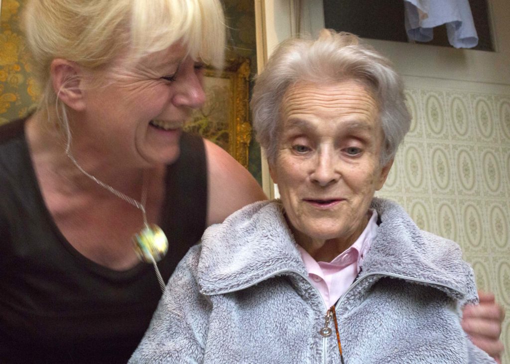 Une auxiliaire de vie sourit à une dame âgée dont elle tient l'épaule, chez elle