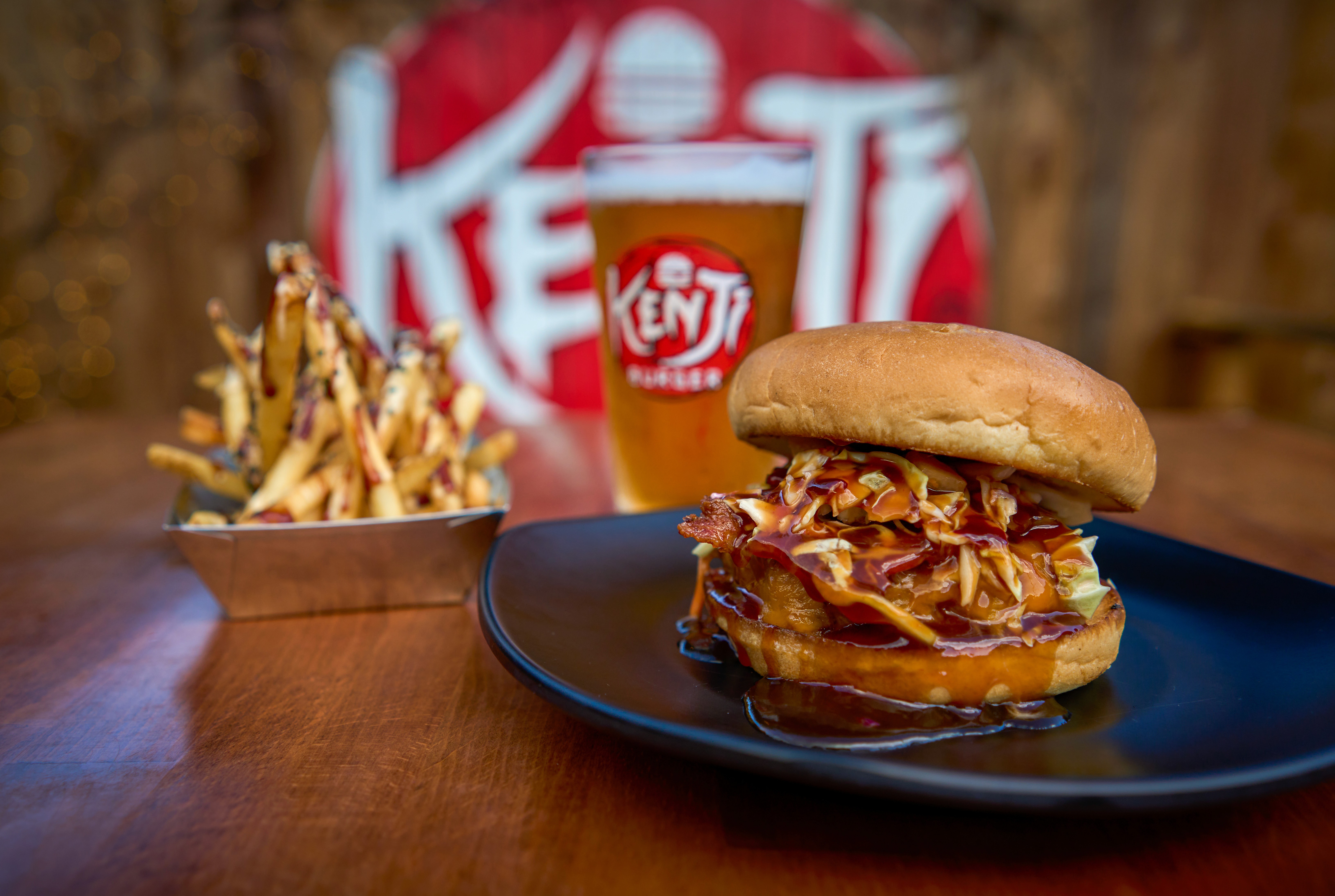 A bright, modern dining room featuring a long light wood table set with several matching wooden chairs and white cushions, illuminated by natural light streaming through large windows overlooking a lush green and sunlit yard.
