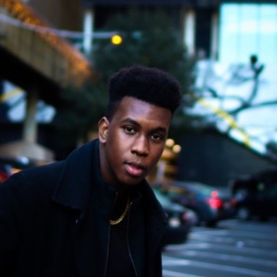 Person in denim shirt over striped tee standing outdoors with blurred buildings in the background