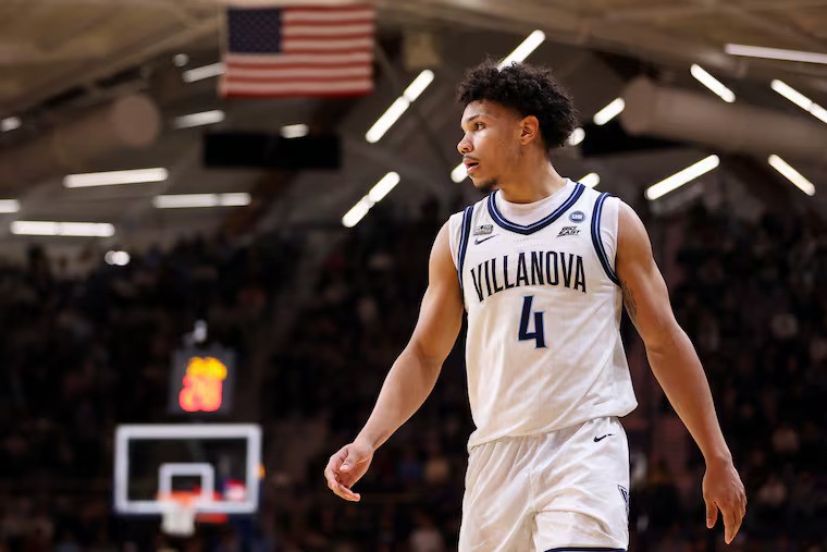 Tyler Perkins in a white Villanova jersey with the number 4 stands on the court, looking to the side. An American flag hangs overhead, and a basketball hoop and scoreboard are visible in the background.
