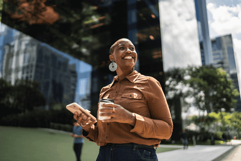 A person stood infront of an office block. They have their phone and a coffee cup in hand, looking with a smile on their face off into the distance
