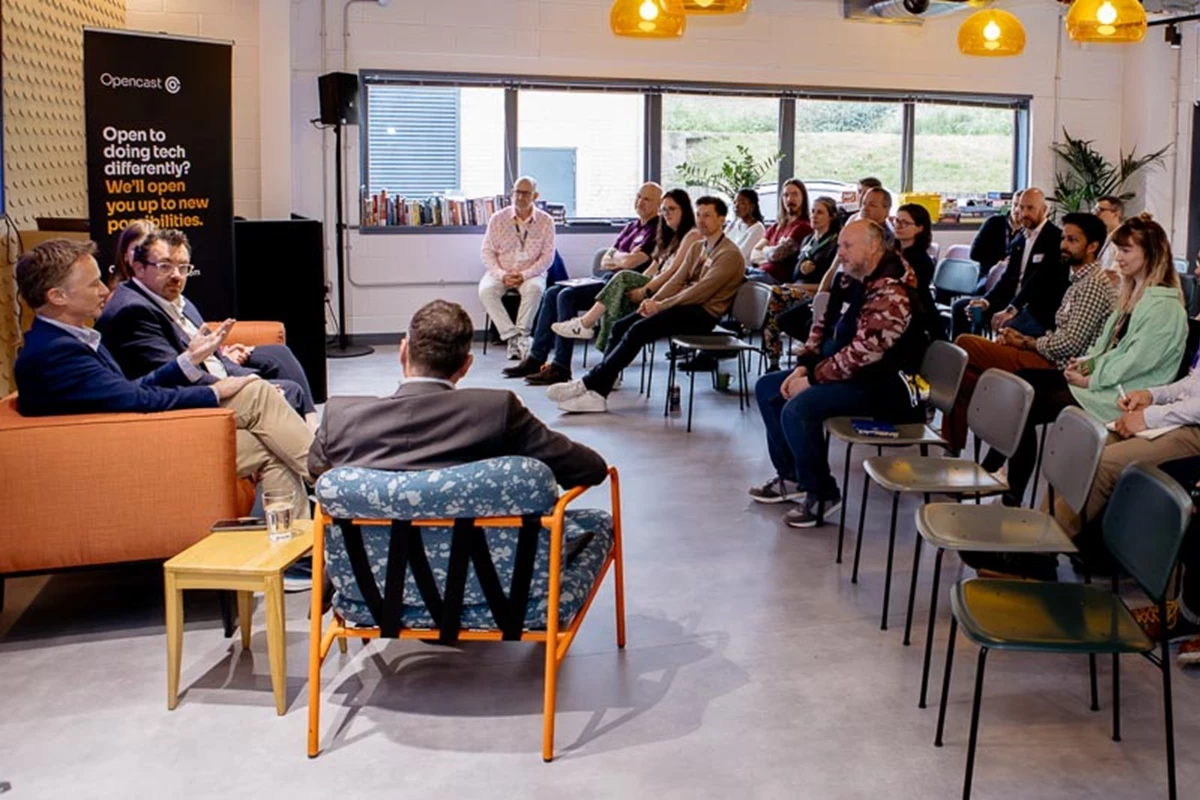 A panel discussion takes place in a modern event space, with speakers seated at the front and an audience arranged in rows of chairs. Warm lighting, large windows, and a banner referencing open technology set a professional atmosphere.