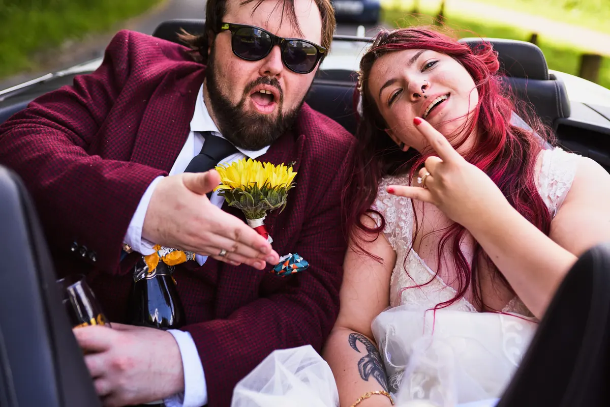 Bride and groom pulling faces in the back of a convertible car