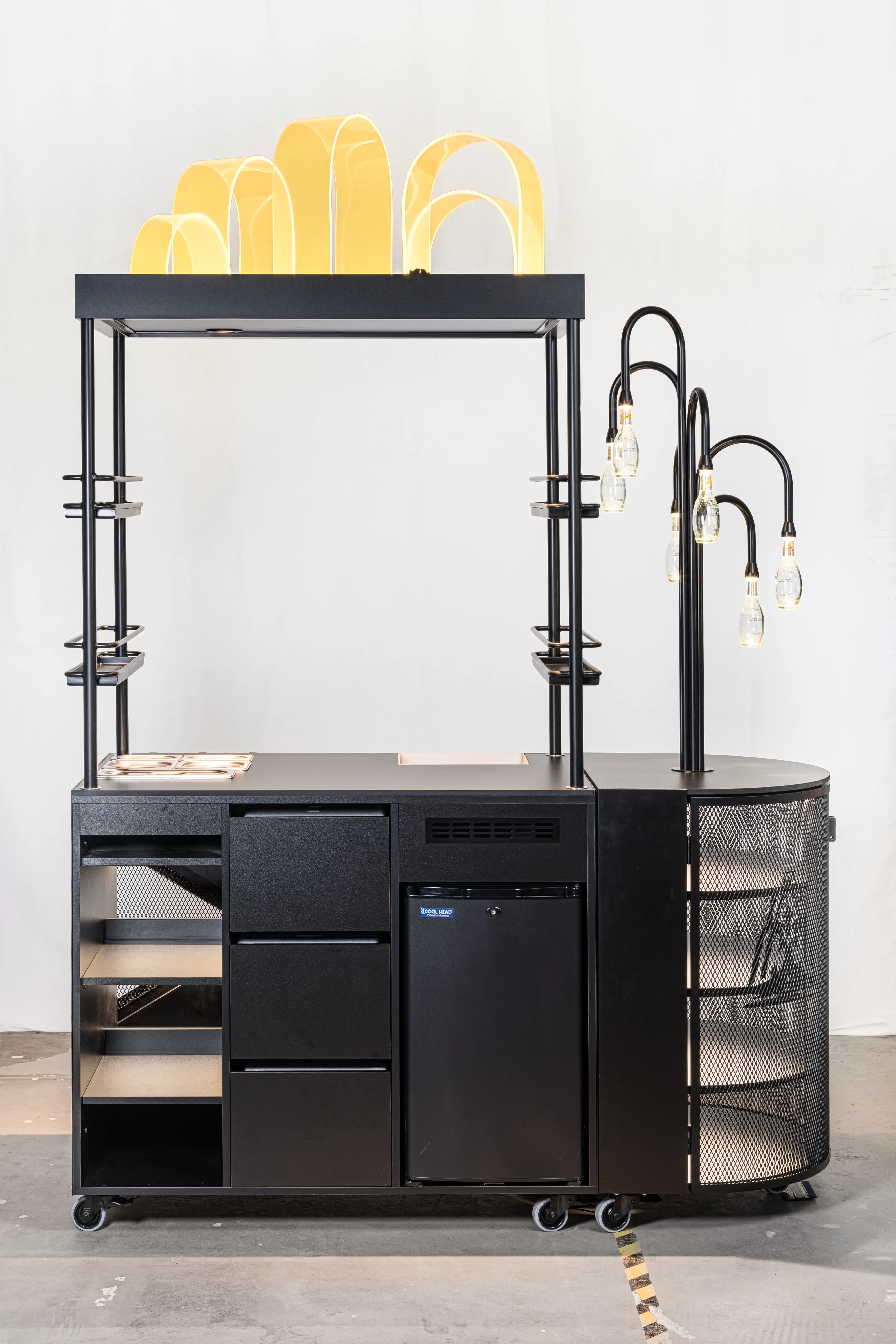Back of the SCHWEPPES bartool with open drawers and shelves, integrated fridge, golden arch decor on top and bottle lamps on a black arm.