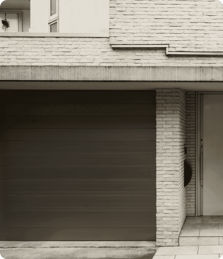 Close-up of a modern, dark bronze or charcoal flush-panel garage door installed under a concrete header. The door is set within a light-colored brick facade and a tiled entryway, featuring a minimalist architectural design with clean horizontal lines.
