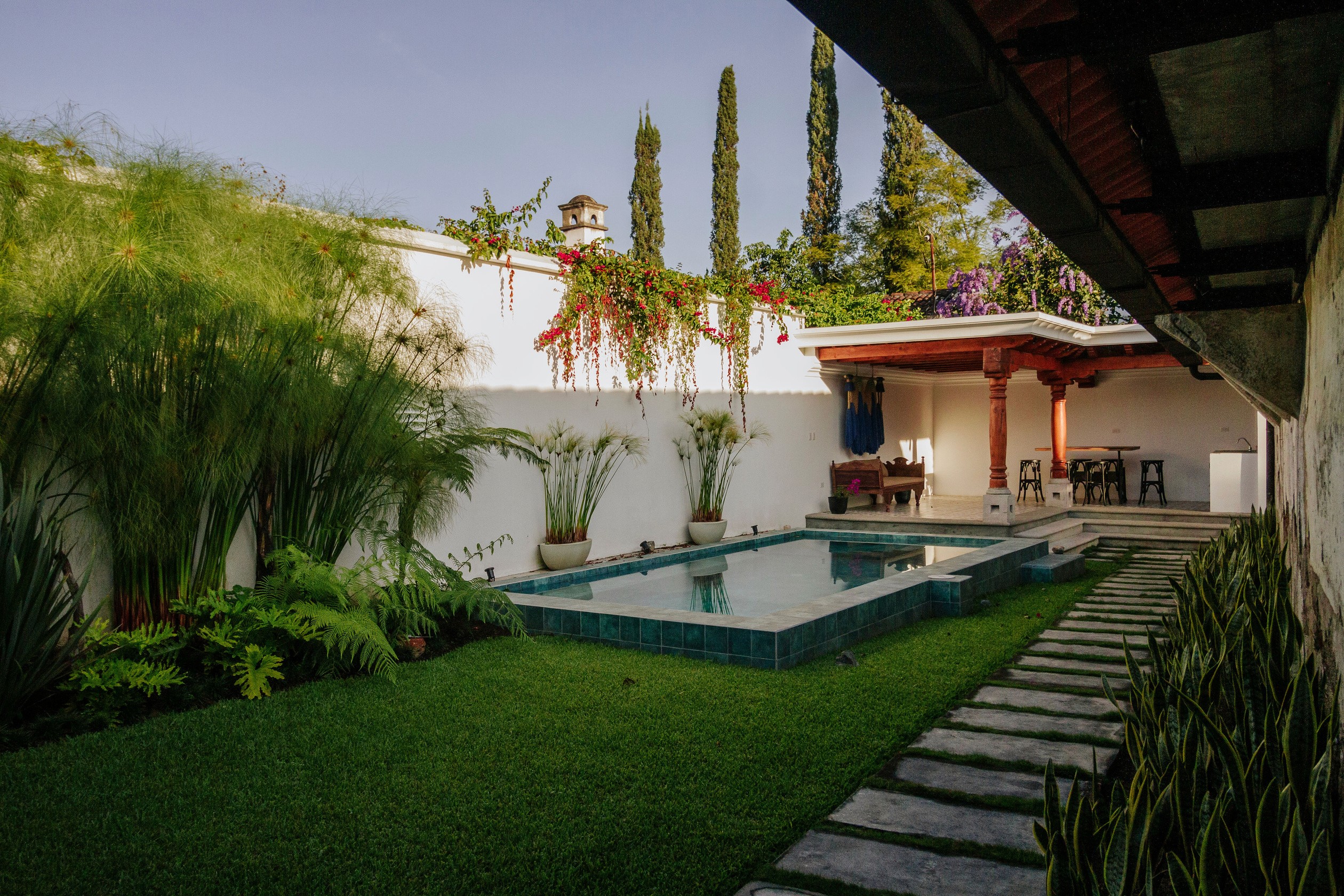 A serene backyard oasis featuring a small, rectangular swimming pool surrounded by lush greenery, stone pathway, and an outdoor seating area under a pergola, set against a white stucco wall adorned with climbing plants.