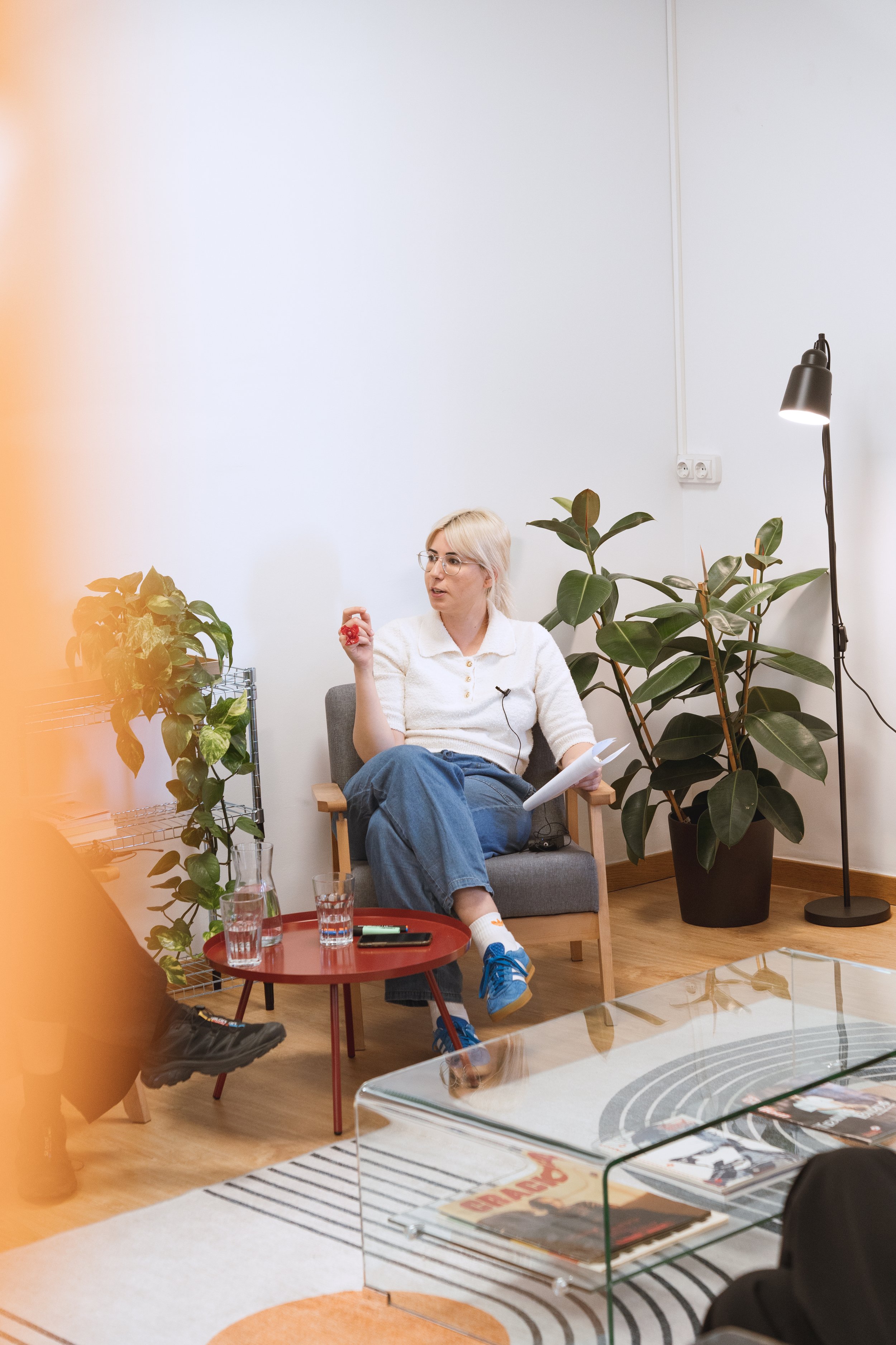 A woman with blonde hair and glasses sitting in a modern living room, holding a red object in one hand and papers in the other. She's seated on a gray armchair next to a red round table with glasses and a phone. There are large green plants nearby and a black floor lamp, with a white wall behind.
