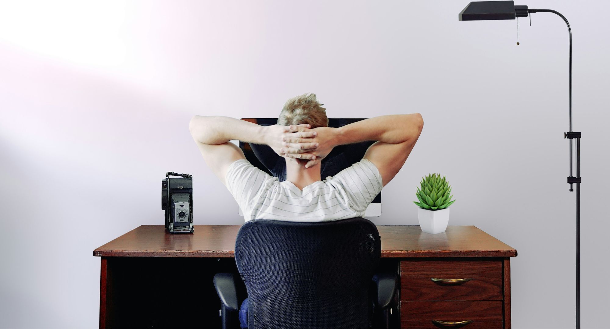 homem de costas relaxado com as mãos na cabeça sentado numa cadeira e uma mesa com computador na frente