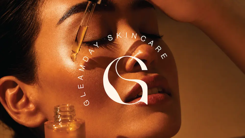 A woman applies a glowing serum on her cheek with a dropper under warm lighting, highlighting radiant and healthy skin, accompanied by a "Gleamday Skincare" logo in the foreground.
