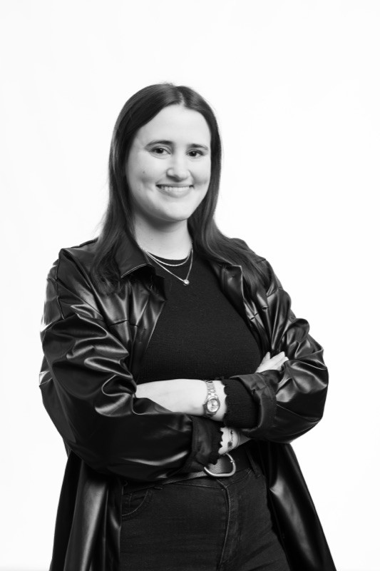 Black-and-white studio portrait of a smiling woman with long straight dark hair, wearing layered necklaces and a wristwatch, in a black oversized leather jacket over a black top, arms crossed, against a plain light background.