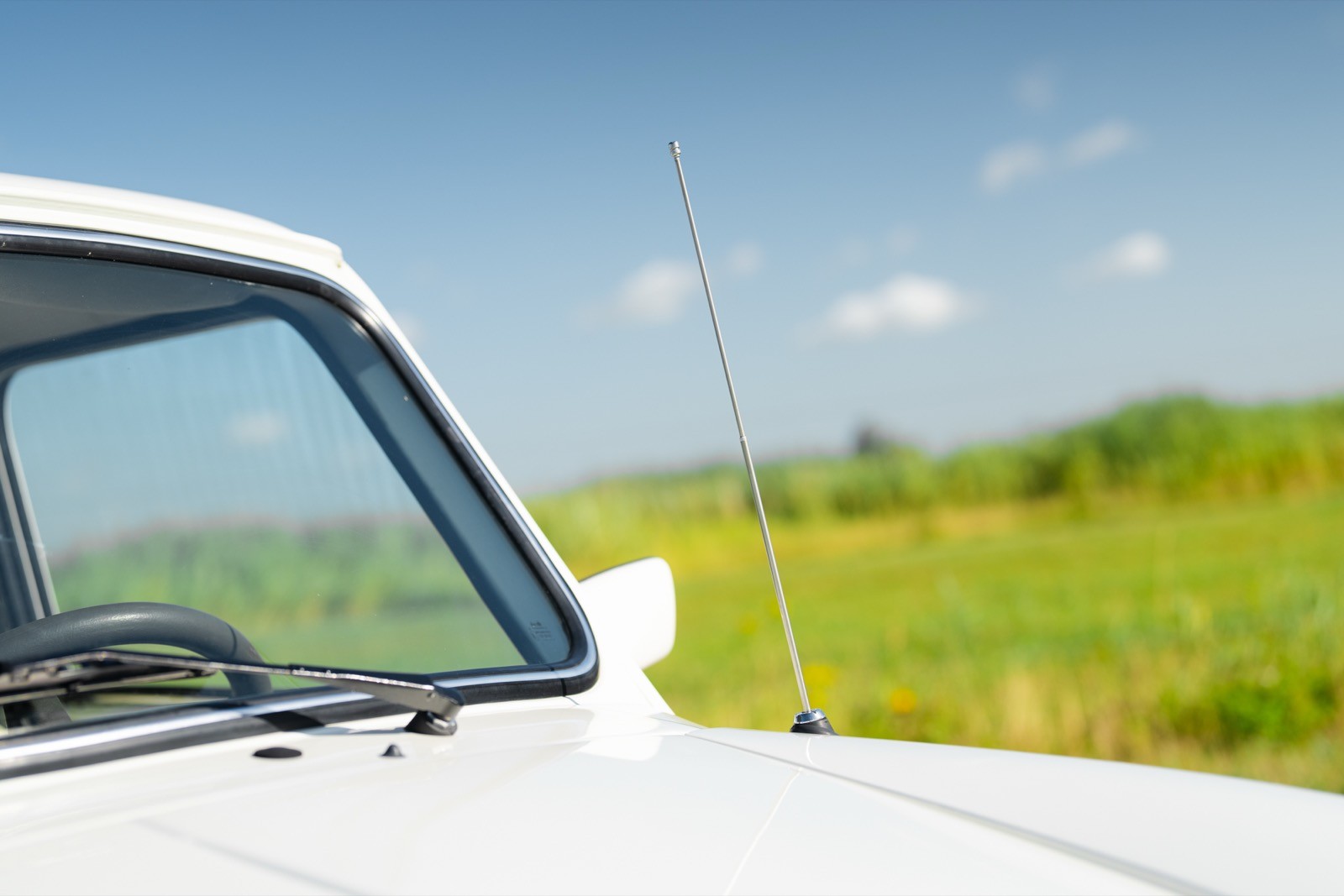 A close-up of a car's side mirror against a background of green fields and a clear blue sky with a few clouds.