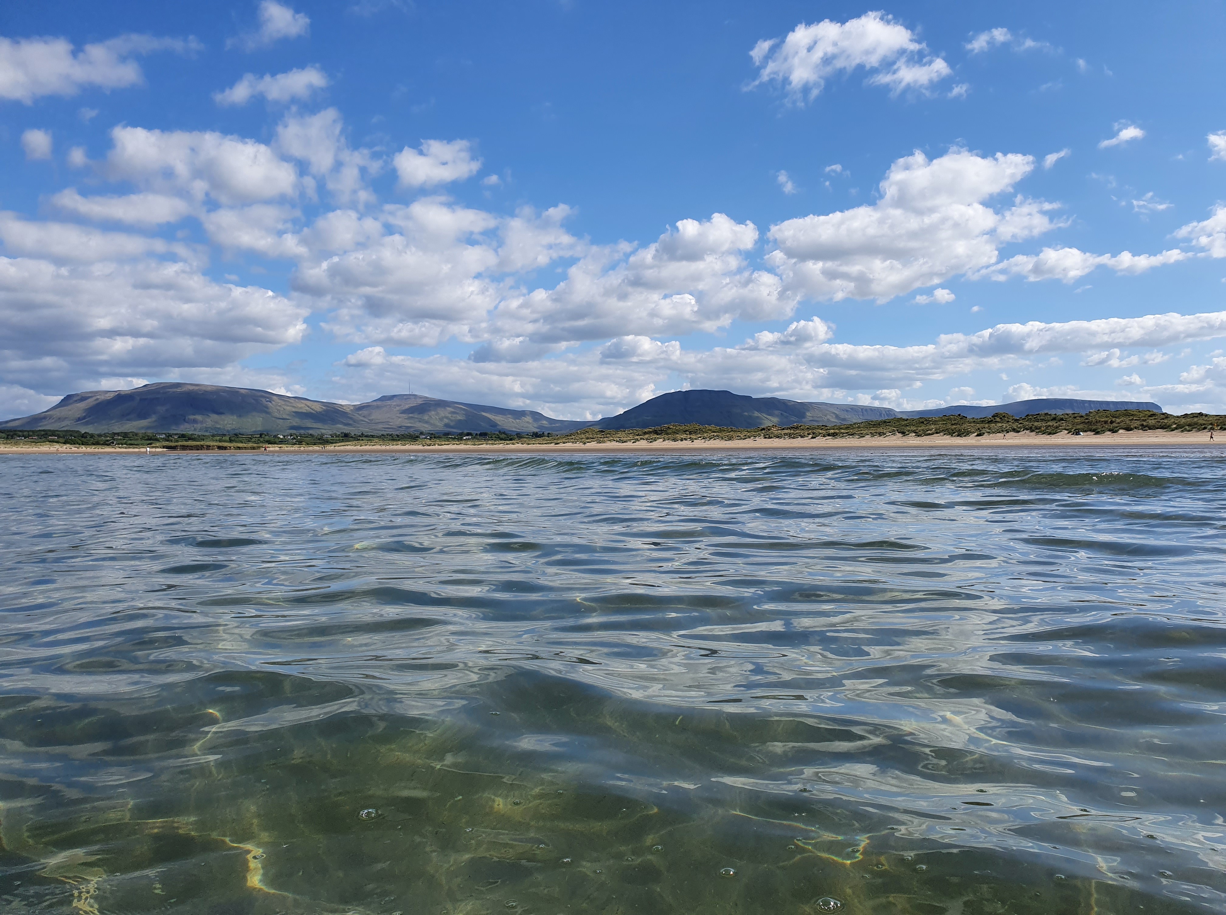 Waves gently lapping at the shore under a bright sky with scattered clouds and distant hills.