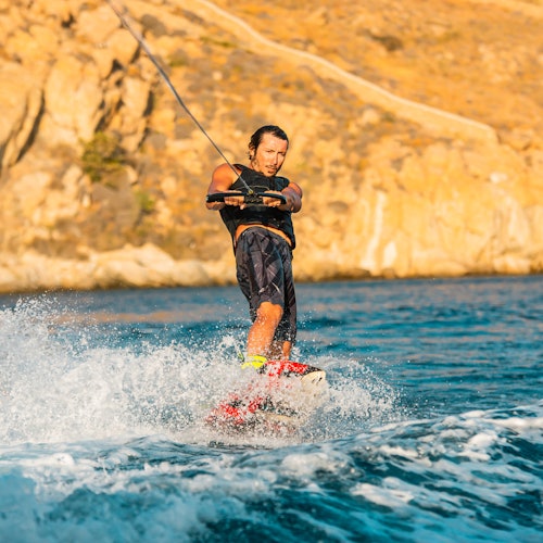 Person wakeboarding on a water body near rocky cliffs, holding a tow rope, with water splashing around them.