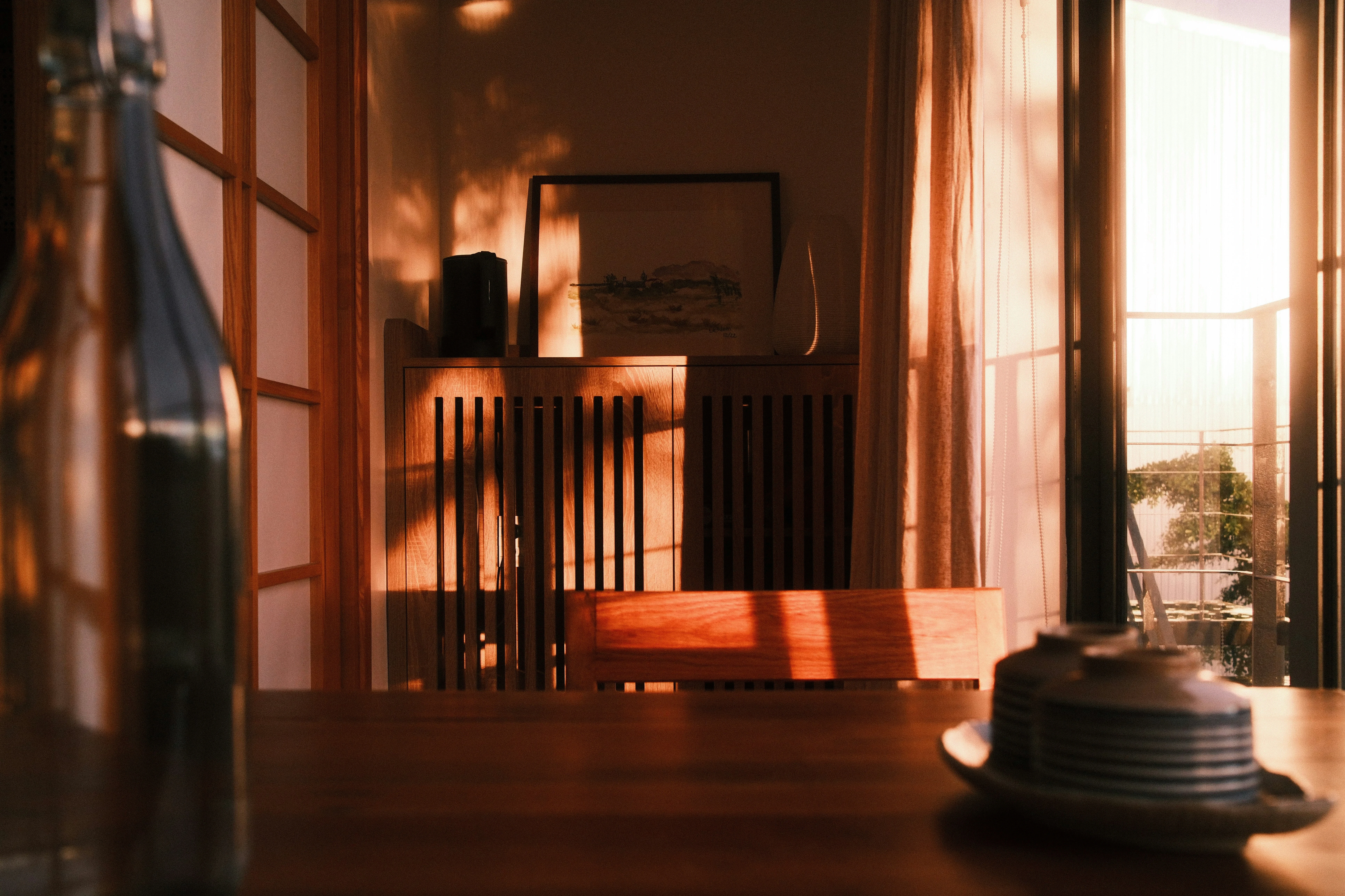 Warm sunset light casts shadows in a cozy room with wooden furniture. A bottle, teacups, and a cabinet are softly illuminated, creating a tranquil atmosphere.