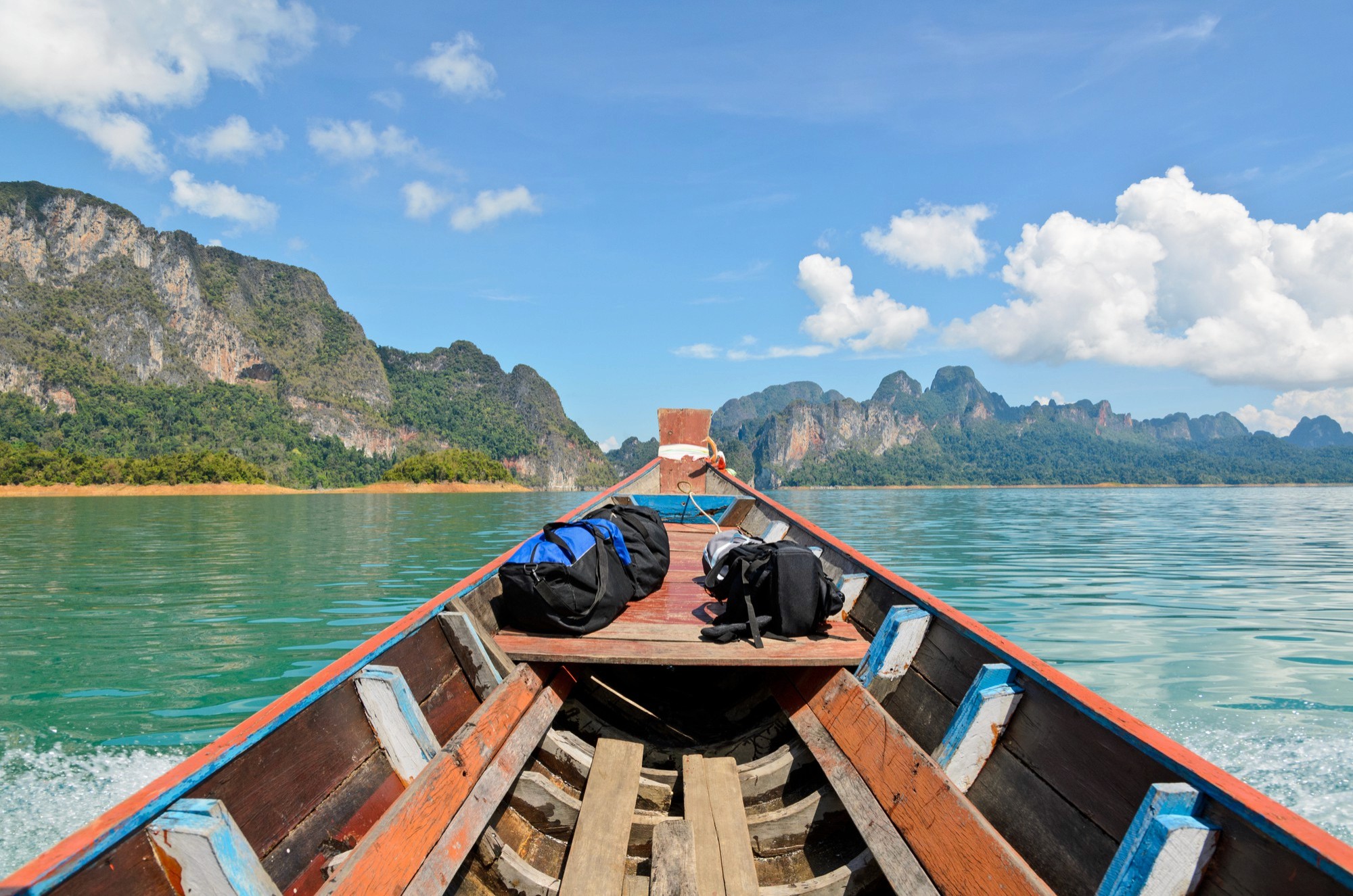 A first-person view from the bow of a traditional wooden longtail boat cruising across a turquoise lake, with travel bags on deck and towering limestone karst mountains in the background under a blue sky.